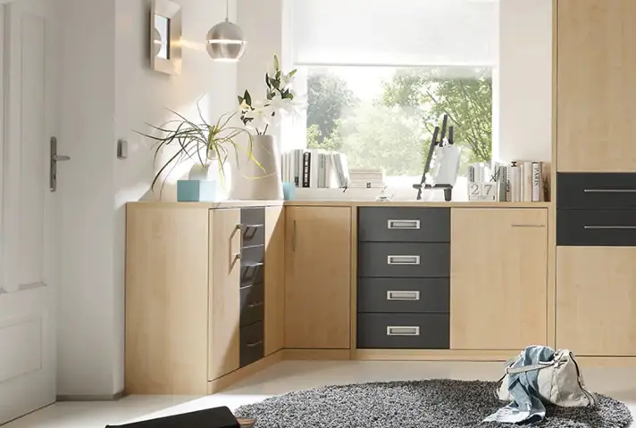 Helles Ecksofa mit grünem Sitzelement und gemusterten Kissen, davor quadratischer Polsterhocker als Couchtisch mit Tablett und Vase, hinten Regal mit Büchern und Pflanzen.